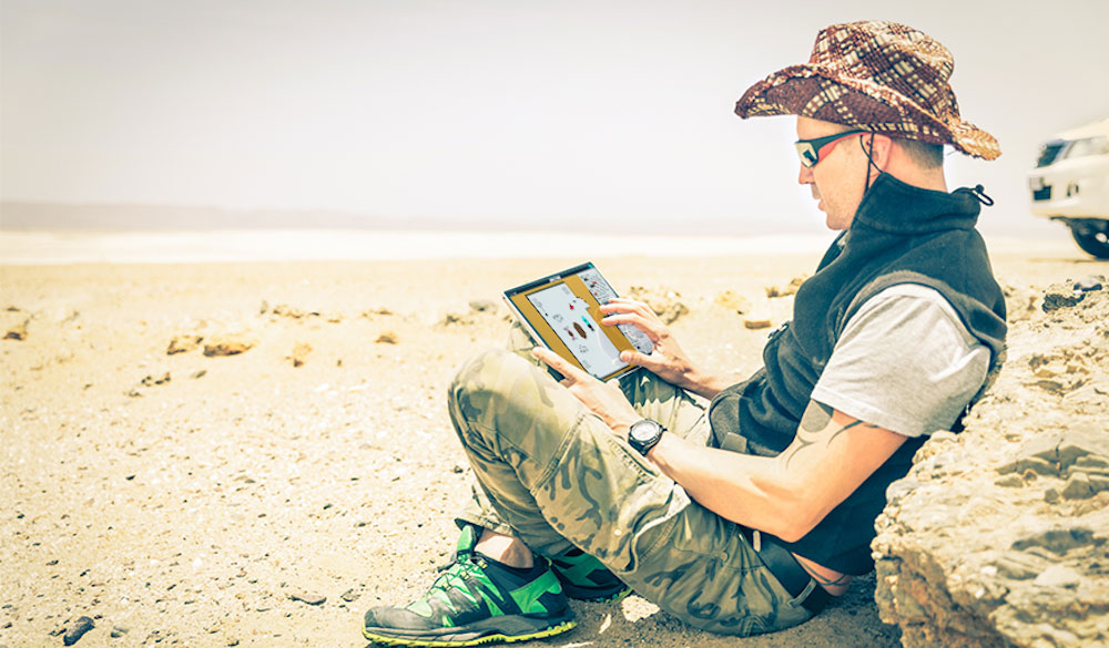 A person wearing camouflage pants sitting against a rock using a laptop.